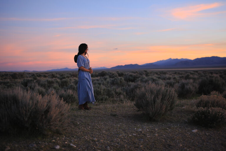 Traditionally dressed girl in long, pale blue dress looks out over a beautiful Nevada sunset. Very old-west feel, traditional American. This is Mercy, she is an American, a mix of almost every race on the planet. The result is stunning.