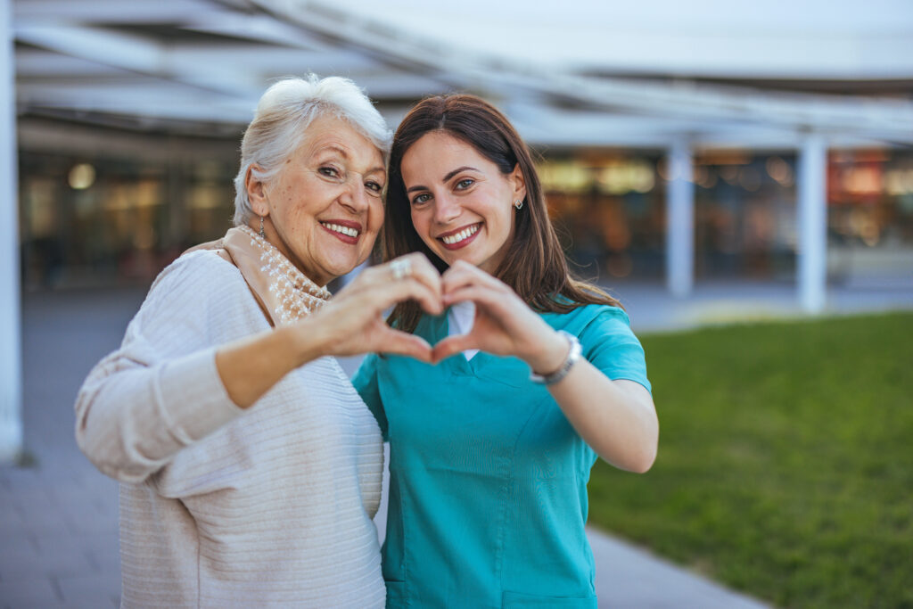 A joyful senior woman and her caregiver make a heart shape with their hands, symbolizing love and support in an outdoor setting.