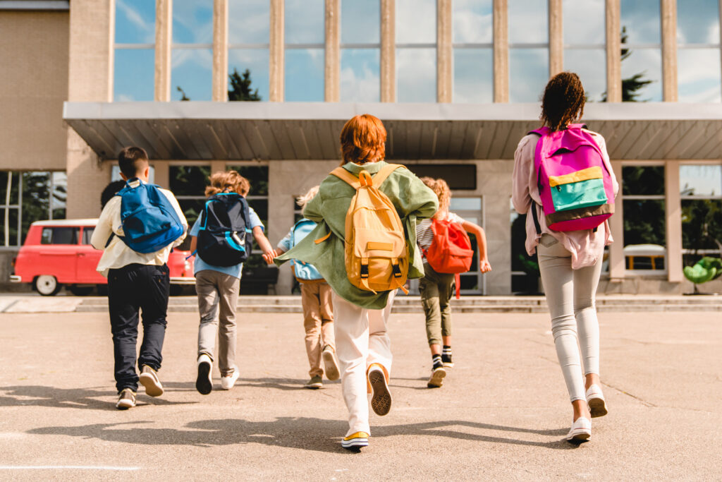 Children with colorful backpacks walking toward a school building.