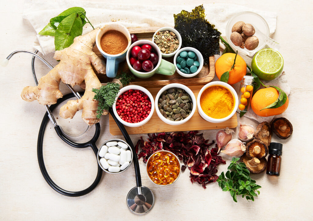 Assortment of herbs, spices, citrus fruits, supplements, and medicinal ingredients arranged around a stethoscope on a wooden surface.
