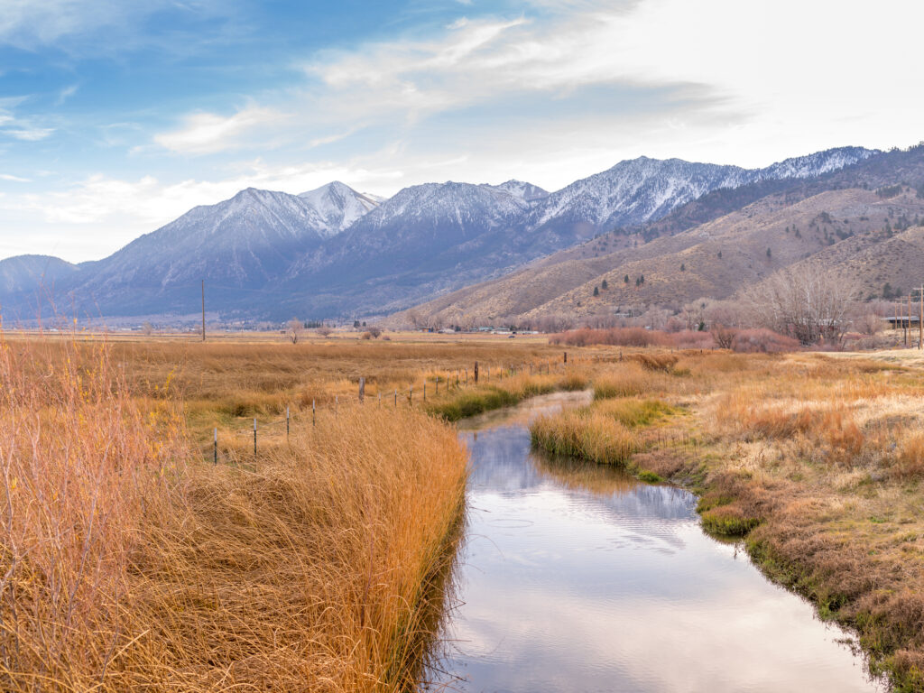 Calm creek bordered by dry fields and fence line, with rugged mountains under a partly cloudy sky.