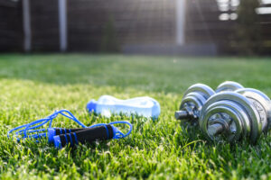 A blue jump rope, a blue water bottle, and silver dumbbells are placed on green grass in a sunny outdoor setting.