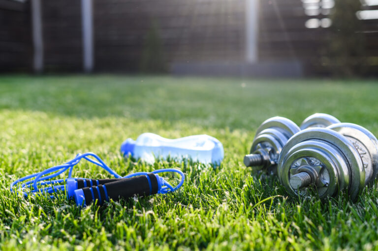 A blue jump rope, a blue water bottle, and silver dumbbells are placed on green grass in a sunny outdoor setting.