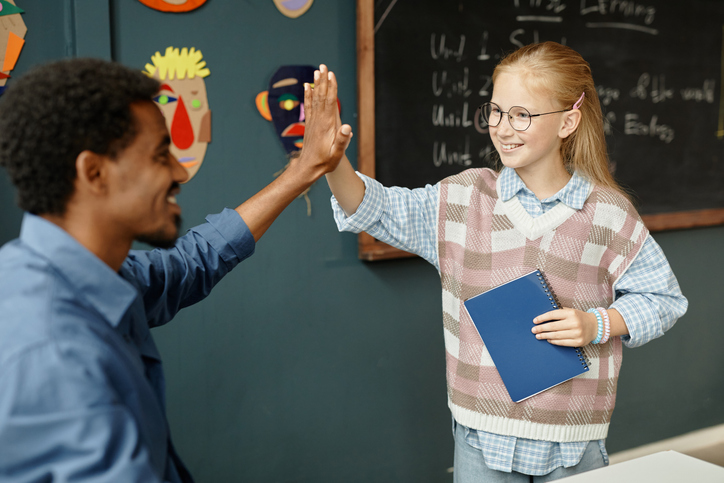 Cheerful young student high-fiving teacher after receiving praise for excellent work during a primary school lesson.