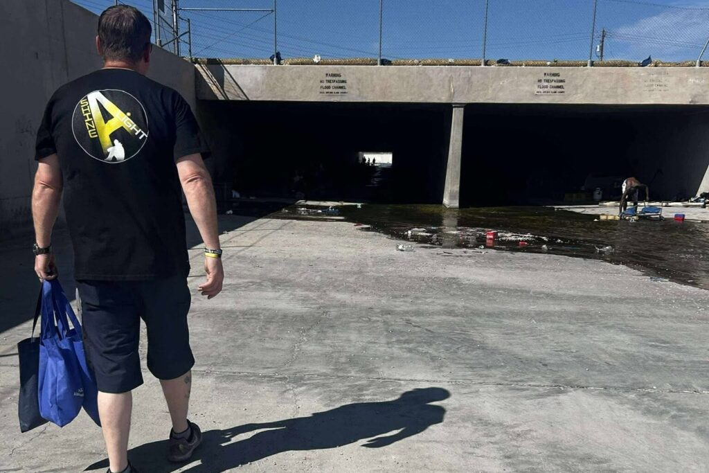 A Shine A Light outreach worker in a branded black shirt walks across a concrete flood channel carrying a blue tote bag, heading toward a shaded tunnel underpass marked with No Trespassing Flood Channel warning signs.