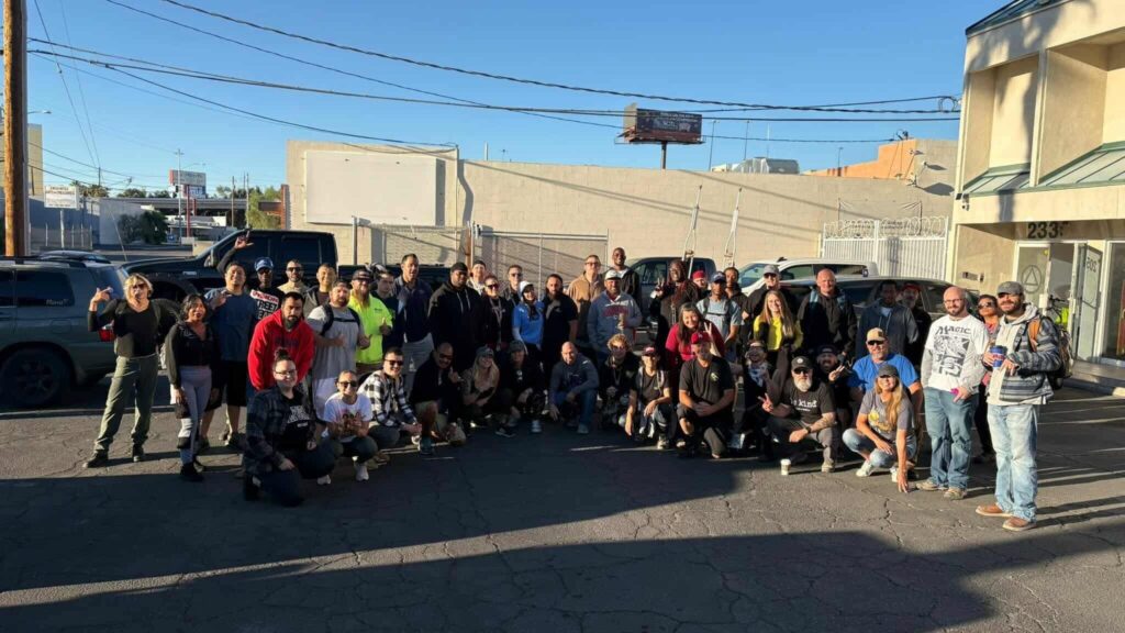 A large group of about 40 Shine A Light outreach volunteers and community members pose together in a Las Vegas parking lot on a sunny morning, many smiling and giving peace signs, with parked cars and a block wall behind them.