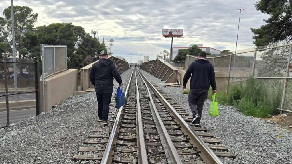 Two Shine A Light outreach workers walk away from the camera along elevated railroad tracks under a cloudy sky, each carrying a colored tote bag of supplies, with Las Vegas buildings and a billboard in the distance.