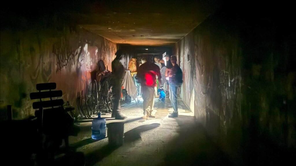 Outreach team members stand inside a dark storm-drain tunnel lit by a single shaft of light, surrounded by bicycles, belongings, and a water jug, as they connect with people living there.