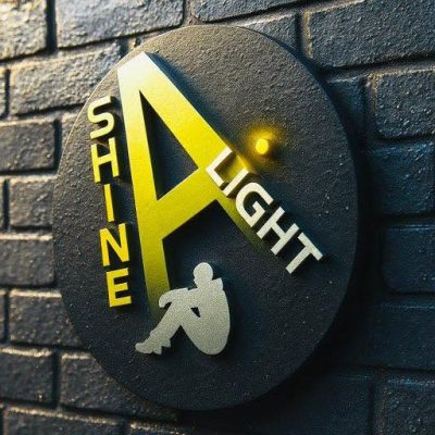 The Shine A Light Foundation logo mounted on a dark brick wall: a circular sign with a yellow letter A shaped like a spotlight beam shining down on a seated figure, and the words Shine A Light around it.