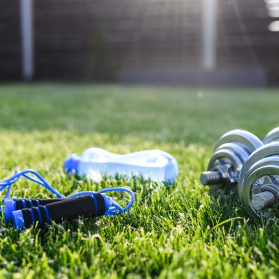 A blue jump rope, a blue water bottle, and silver dumbbells are placed on green grass in a sunny outdoor setting.