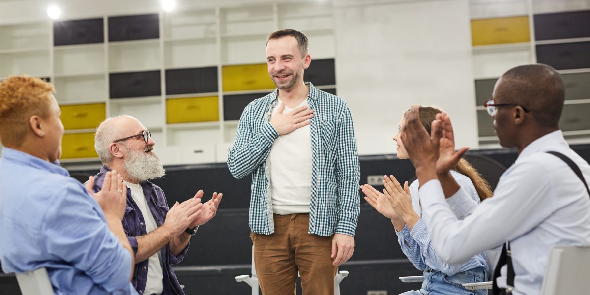 Portrait of smiling mature man introducing himself during therapy session in support group to people clapping, copy space