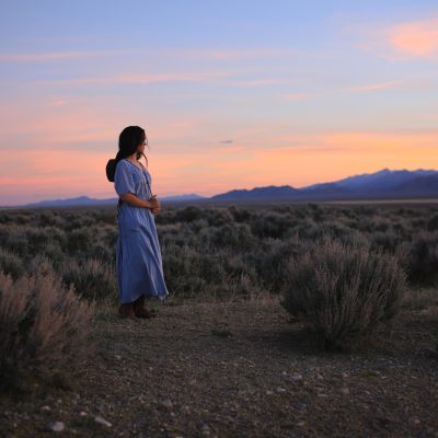 Traditionally dressed girl in long, pale blue dress looks out over a beautiful Nevada sunset. Very old-west feel, traditional American. This is Mercy, she is an American, a mix of almost every race on the planet. The result is stunning.