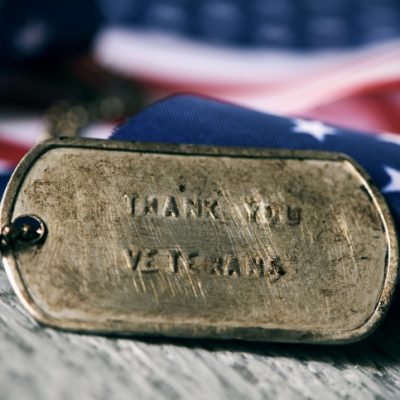 text thank you veterans in a dog tag closeup of a rusty dog tag with the text thank you veterans engraved in it, next to a flag of the United States, on a rustic wooden surface